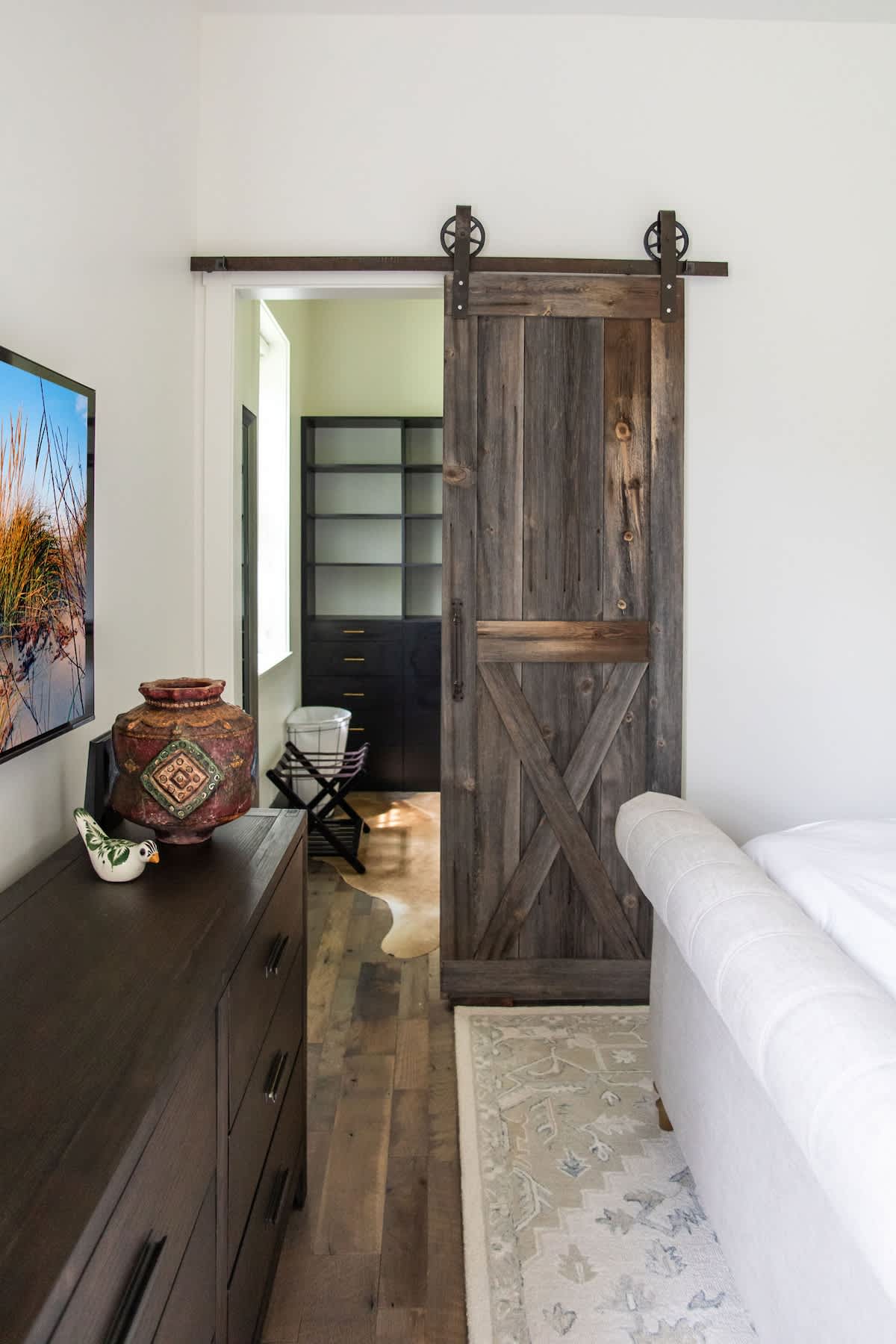 A rustic bedroom featuring a dark wood dresser with decorative pottery and a large TV, with a sliding barn door made of reclaimed wood leading to a walk-in closet. Hardwood floors and a white sofa are also visible.