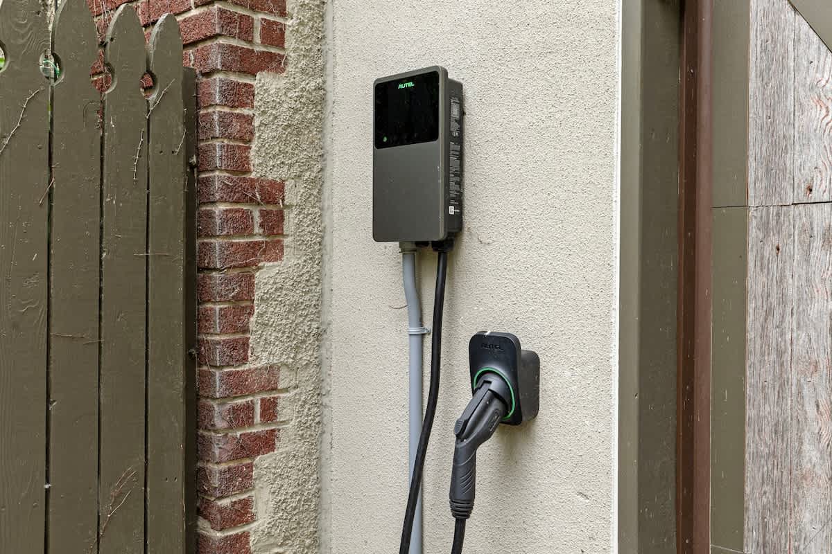 A dark gray EV charger is mounted on a light-colored stucco wall, with its charging cable neatly coiled, positioned next to a rustic wooden gate and a brick wall.