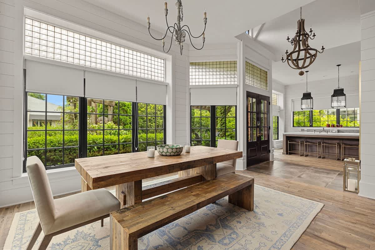 Open-concept view showing a bright dining room with a rustic wooden table and bench, leading into a kitchen area visible through an archway. The space features large black-framed windows, textured glass transoms, and white shiplap walls.