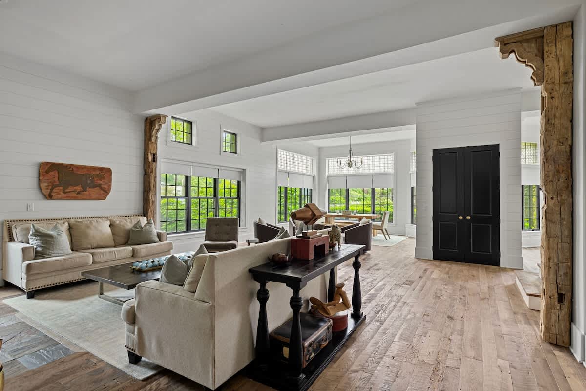 Bright and airy living room with two light-colored sofas, a dark console table behind one, and large windows providing ample natural light. Rustic wooden columns and shiplap walls enhance the modern farmhouse aesthetic.