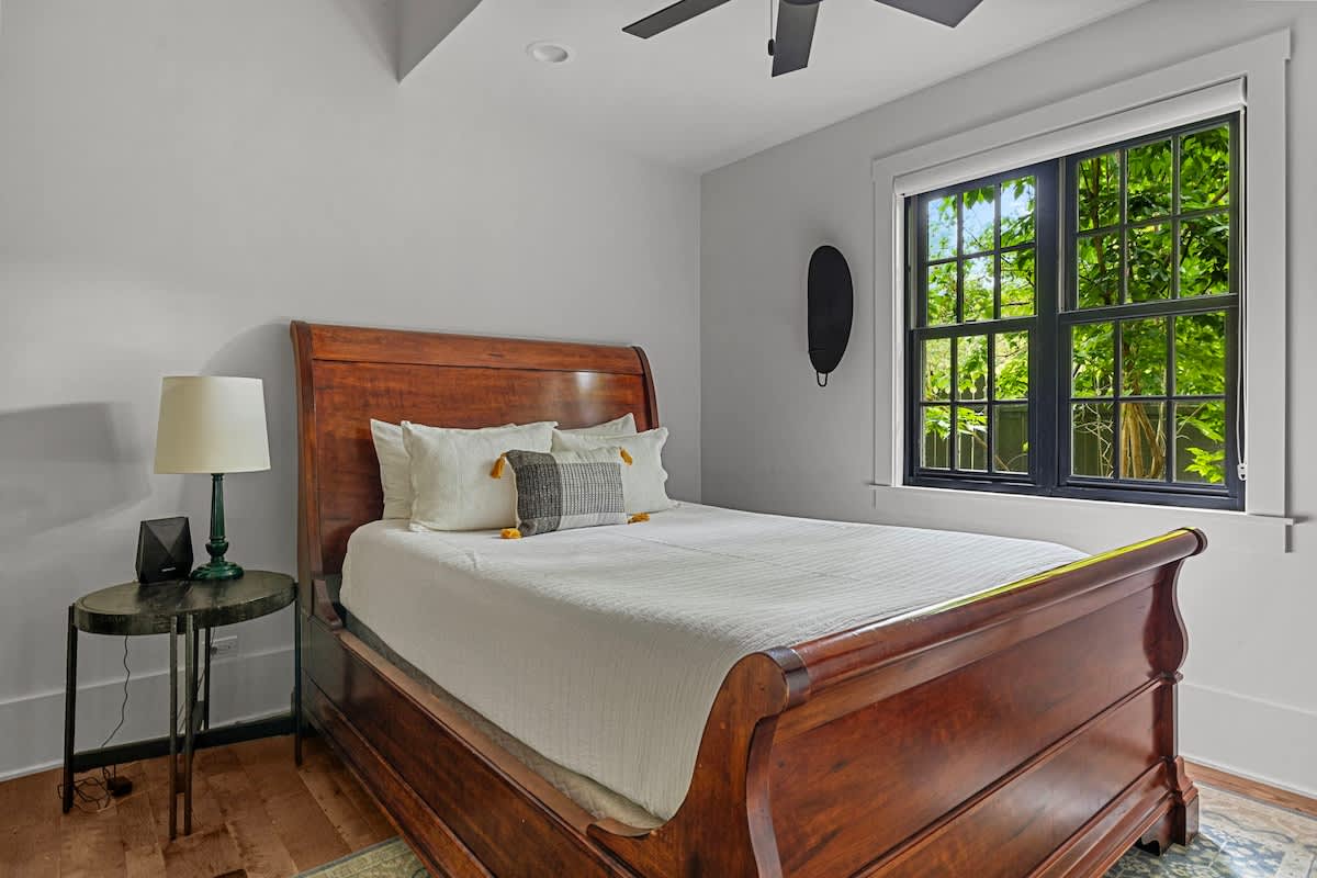 A cozy bedroom features a rich wooden sleigh bed with white bedding and decorative pillows, flanked by a dark nightstand with a green lamp. A large black-framed window offers a vibrant view of lush green trees outside.