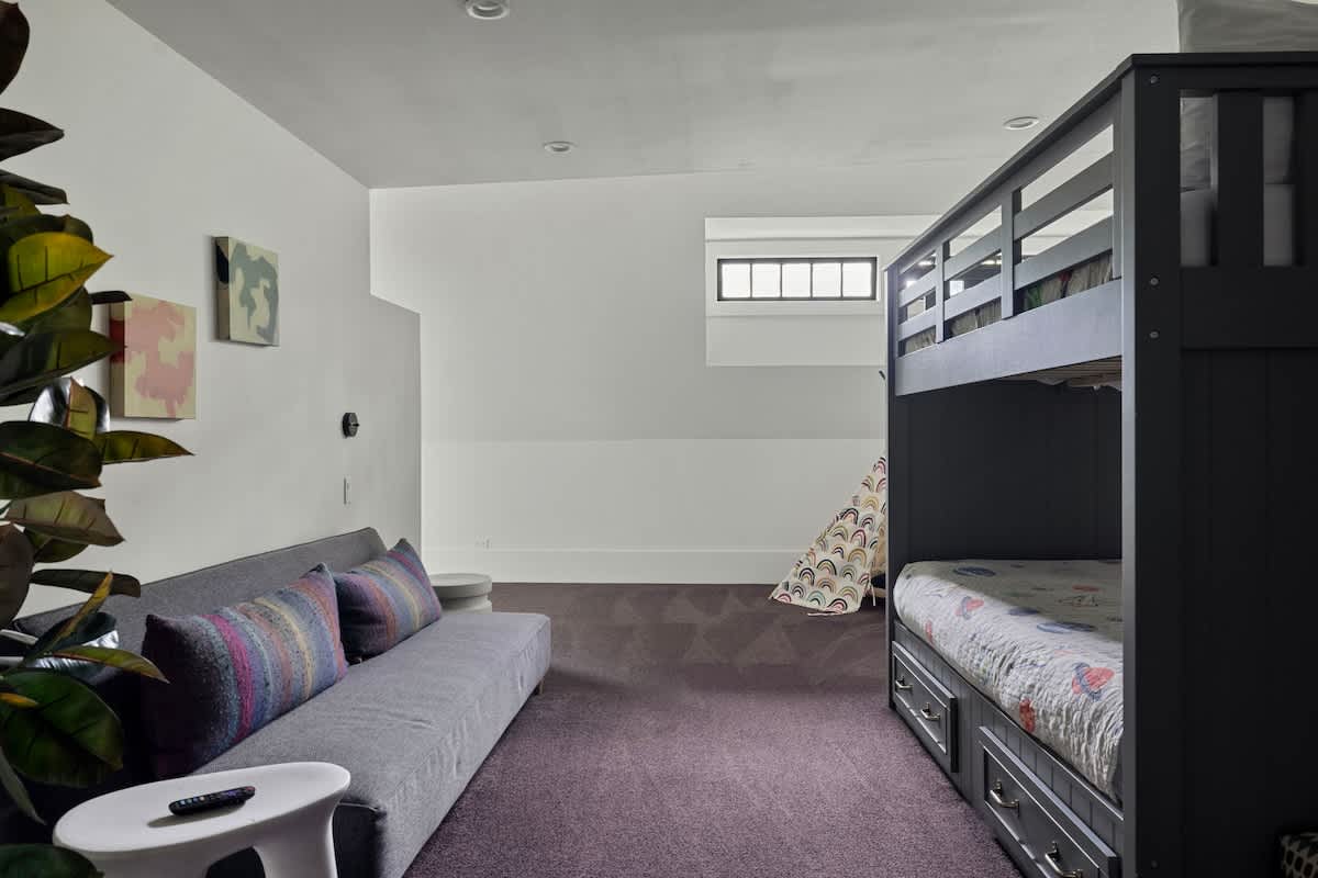 A spacious kids' room with a grey bunk bed and a matching grey sofa with colorful pillows, set against white walls and purple carpet. A large plant adds greenery, and a small window is visible in the background.