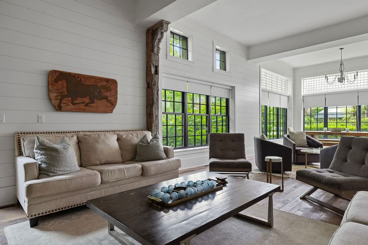 Comfortable living room with a light-colored sofa, various armchairs, and a dark coffee table with decorative accents. Large windows and shiplap walls contribute to a bright and relaxed atmosphere.