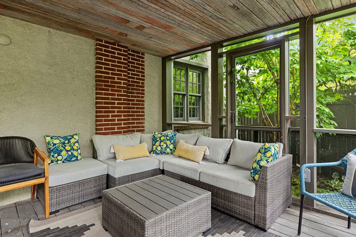 Screened-in porch featuring a comfortable wicker sectional sofa with light cushions and colorful throw pillows, a matching coffee table, and a view of lush greenery.