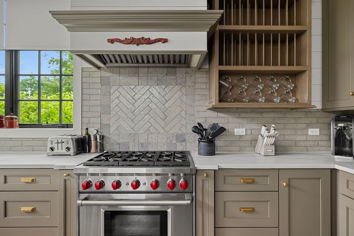 Close-up of a kitchen area featuring a stainless steel range with red knobs, set against a light gray subway tile backsplash with a herringbone pattern above the range. Gray shaker cabinets and white marble countertops flank the appliance.