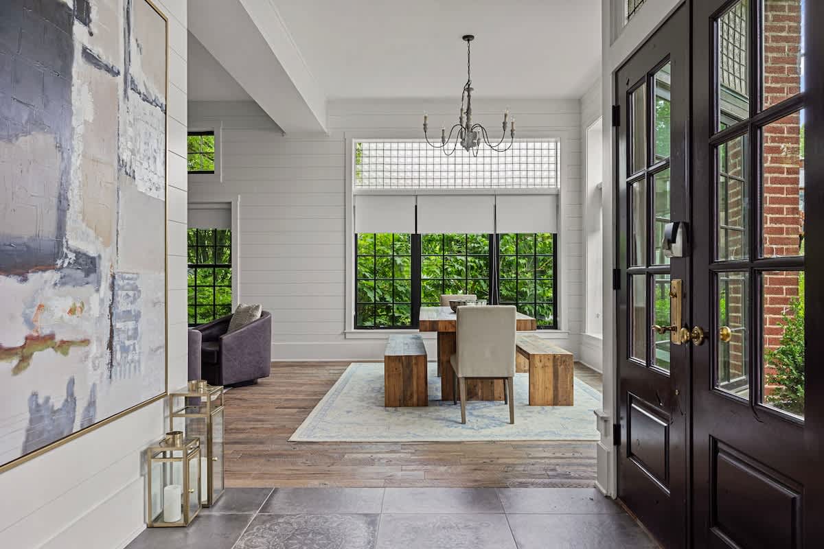 Welcoming entryway with a dark paneled front door and tiled flooring, opening into an open-concept space with a dining area featuring a rustic wooden table and large windows. A large abstract painting adorns the wall.