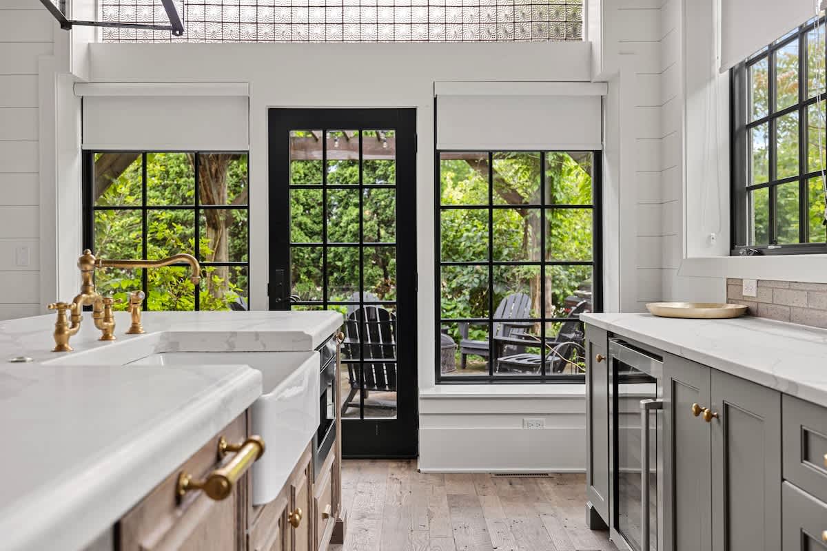View of a kitchen island with a farmhouse sink and brass faucet, facing large black-framed windows and a glass-paned door leading outside. White shiplap walls and textured glass transoms enhance the bright, airy feel.