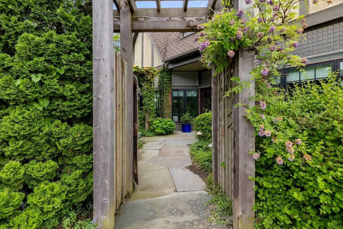 Inviting garden pathway made of flagstones, leading through a rustic wooden gate covered in climbing vines and surrounded by vibrant green foliage towards the house entrance.