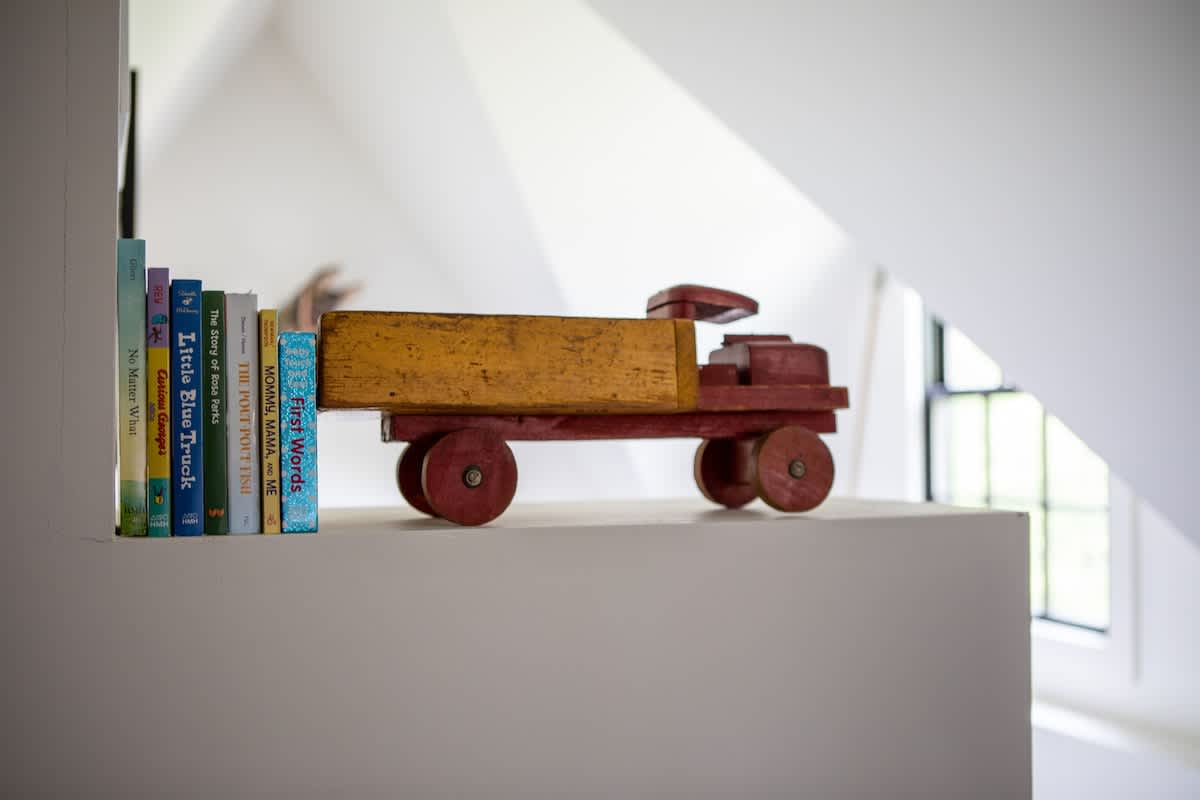 A close-up of a white shelf displaying a collection of children's books and a vintage-style wooden toy truck in red and yellow. The background shows a sloped white wall and a window.