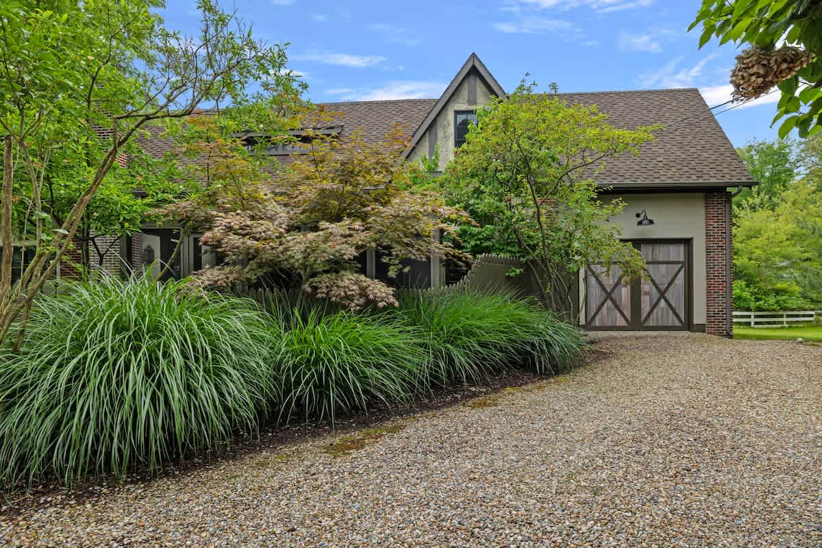 A gravel driveway leads to the front of a house featuring a gabled roof, a rustic wooden garage door with an X-pattern, and lush green landscaping including tall ornamental grasses.