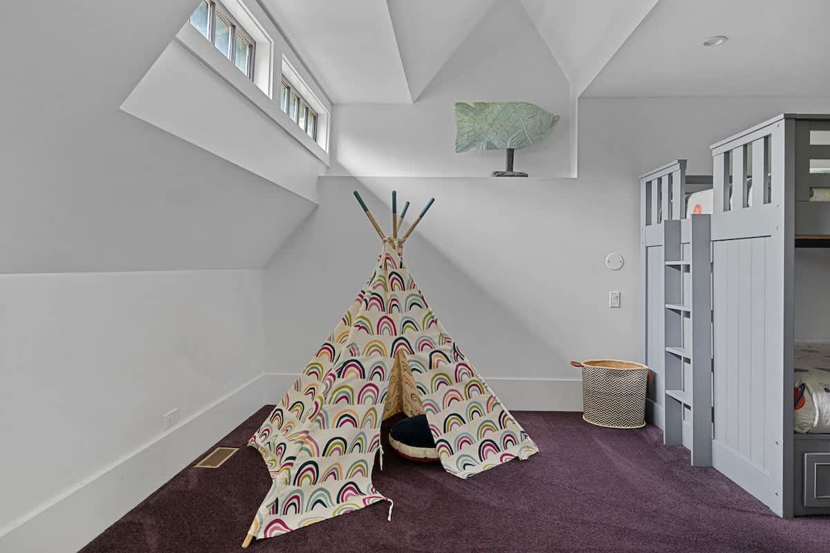 A playful kids' room in an attic space with sloped white walls and purple carpet, featuring a colorful teepee and a grey bunk bed. A decorative fish sculpture hangs on the wall, and high windows let in light.
