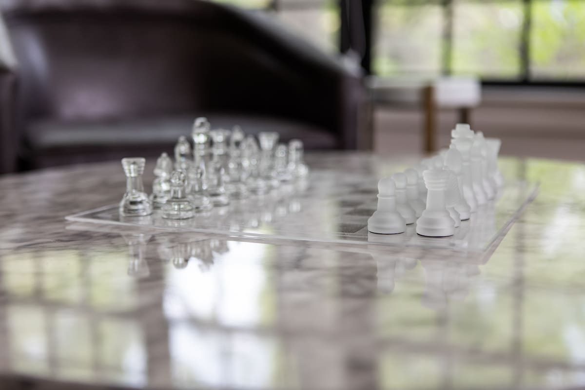 Elegant glass chess set displayed on a polished marble coffee table, with a blurred background showing a living area with a dark armchair.