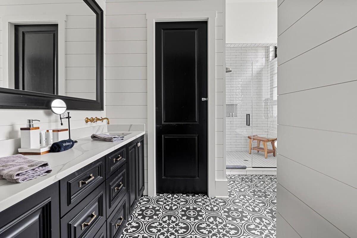 A modern farmhouse-style bathroom featuring a black double vanity with a white countertop, shiplap walls, and striking black and white patterned floor tiles. A black door and a glass-enclosed shower with subway tiles are visible in the background.