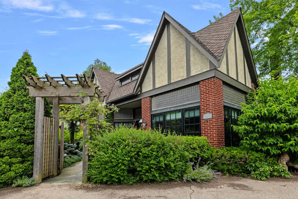 The front exterior of a Tudor-style house features a steep gabled roof, a facade of brick and stucco, and large windows, with a wooden pergola entrance framed by dense green landscaping.