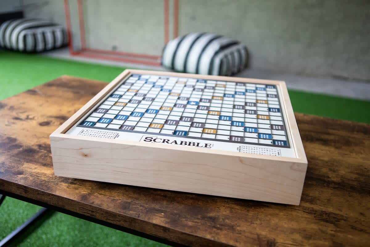 Close-up of a classic Scrabble board game set on a rustic wooden coffee table, with soft poufs visible in the background on artificial turf.