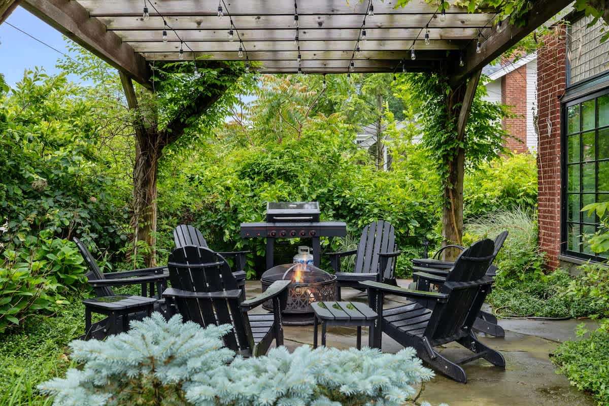 Charming outdoor patio area under a wooden pergola, featuring black Adirondack chairs arranged around a fire pit, with a BBQ grill nearby, all nestled within dense greenery.