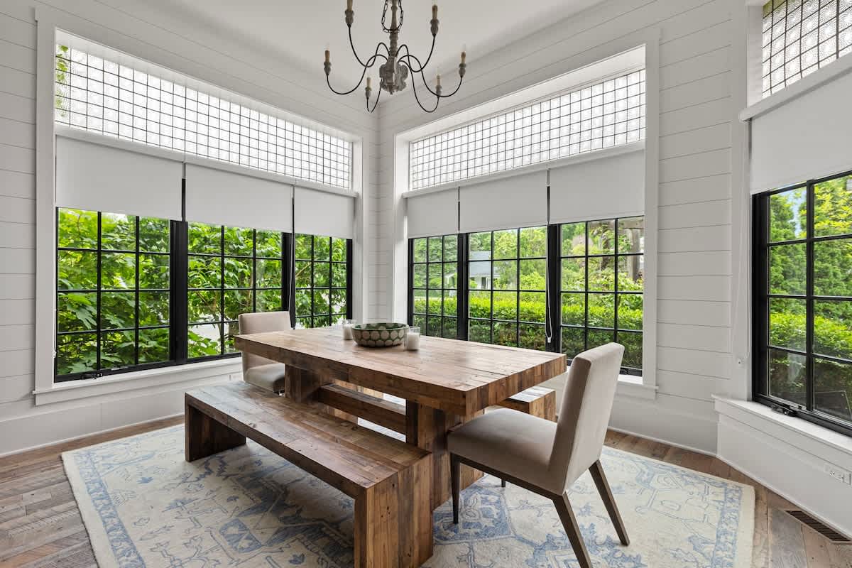 Bright dining room with a rustic wooden table and bench, accompanied by two upholstered chairs, all resting on a light blue patterned rug. Large black-framed windows and textured glass transoms flood the room with natural light, set against white shiplap walls.