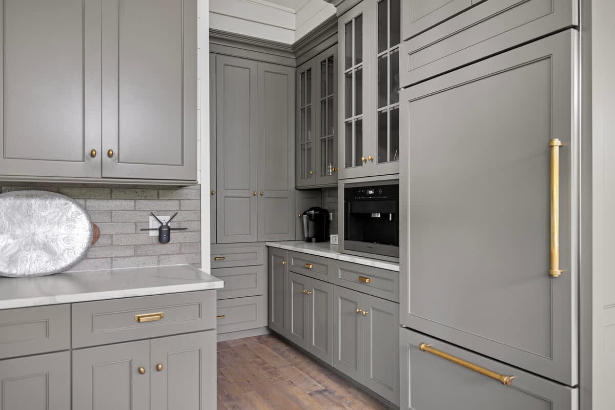 Section of a modern kitchen featuring gray shaker cabinets with brass hardware, a built-in coffee maker, and a panel-ready refrigerator. White marble countertops and a light gray brick backsplash complete the sophisticated look.
