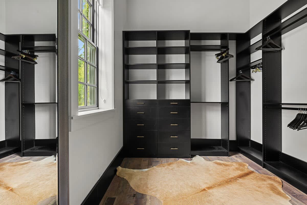 A spacious walk-in closet features sleek black shelving units, drawers with gold pulls, and ample hanging space. A large window provides natural light, and a light brown cowhide rug adds a touch of texture to the hardwood floor.