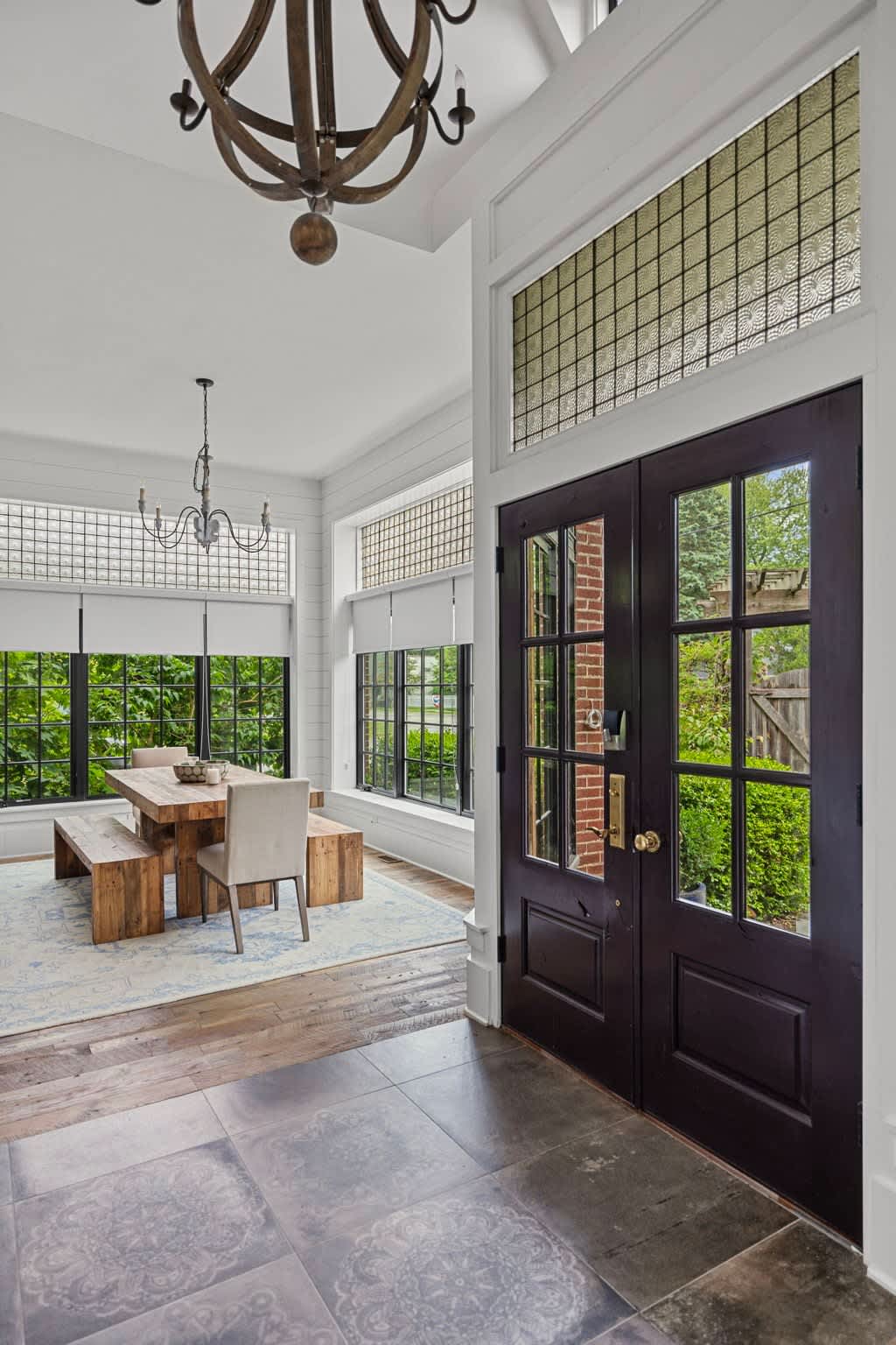 Grand foyer with dark double doors featuring glass panes, leading into a dining area with a rustic wooden table and bench. The space features dark tile flooring, white shiplap walls, and transom windows with textured glass.