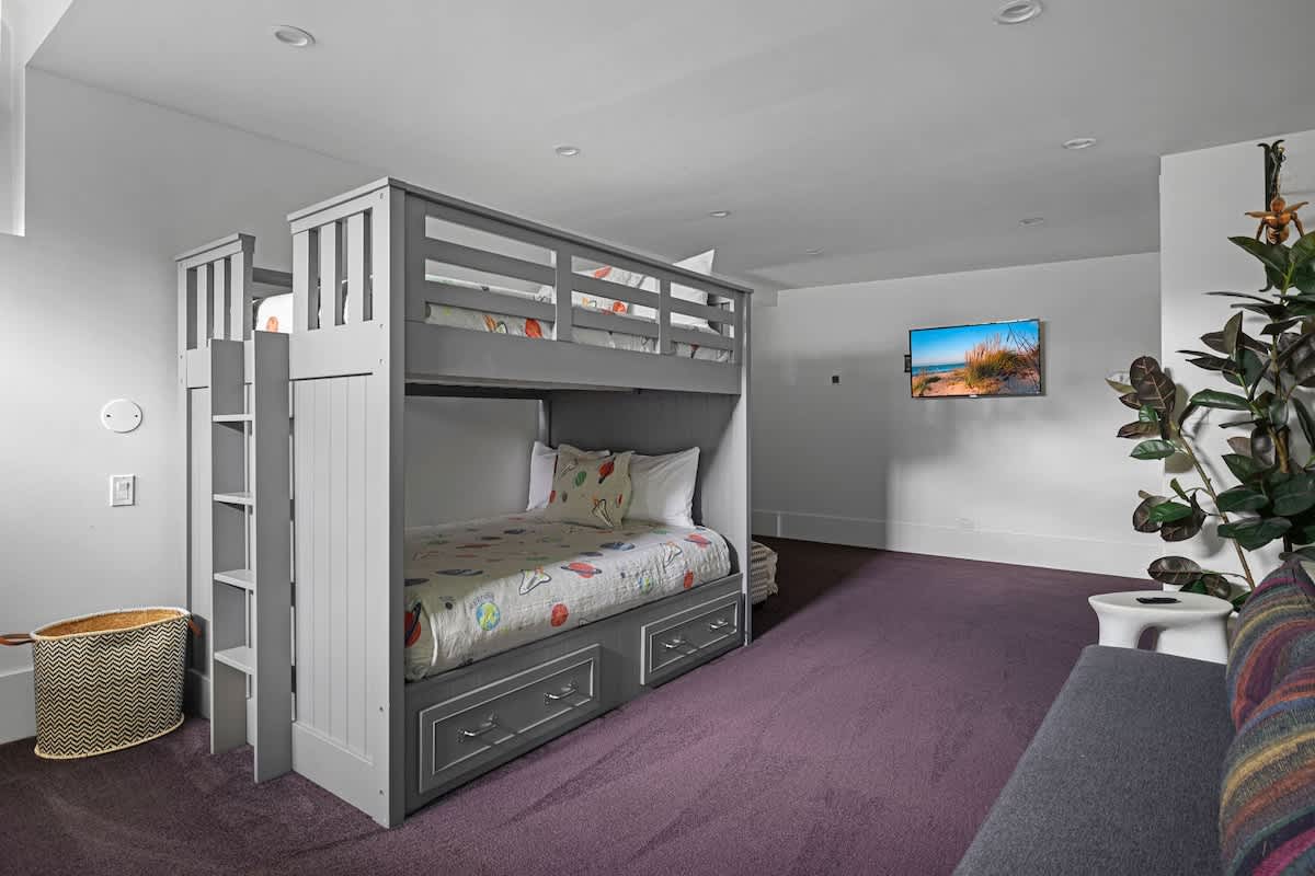 Spacious room with gray bunk beds featuring built-in drawers, a wall-mounted TV displaying a beach scene, and soft purple carpet, ideal for children.