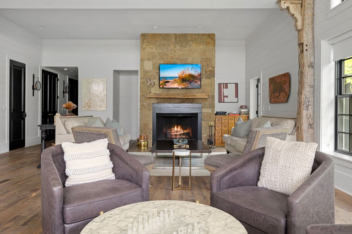 Spacious living room centered around a stone fireplace with a TV mounted above, flanked by two light-colored sofas and two dark swivel chairs. Hardwood floors and white walls create a bright, inviting atmosphere.