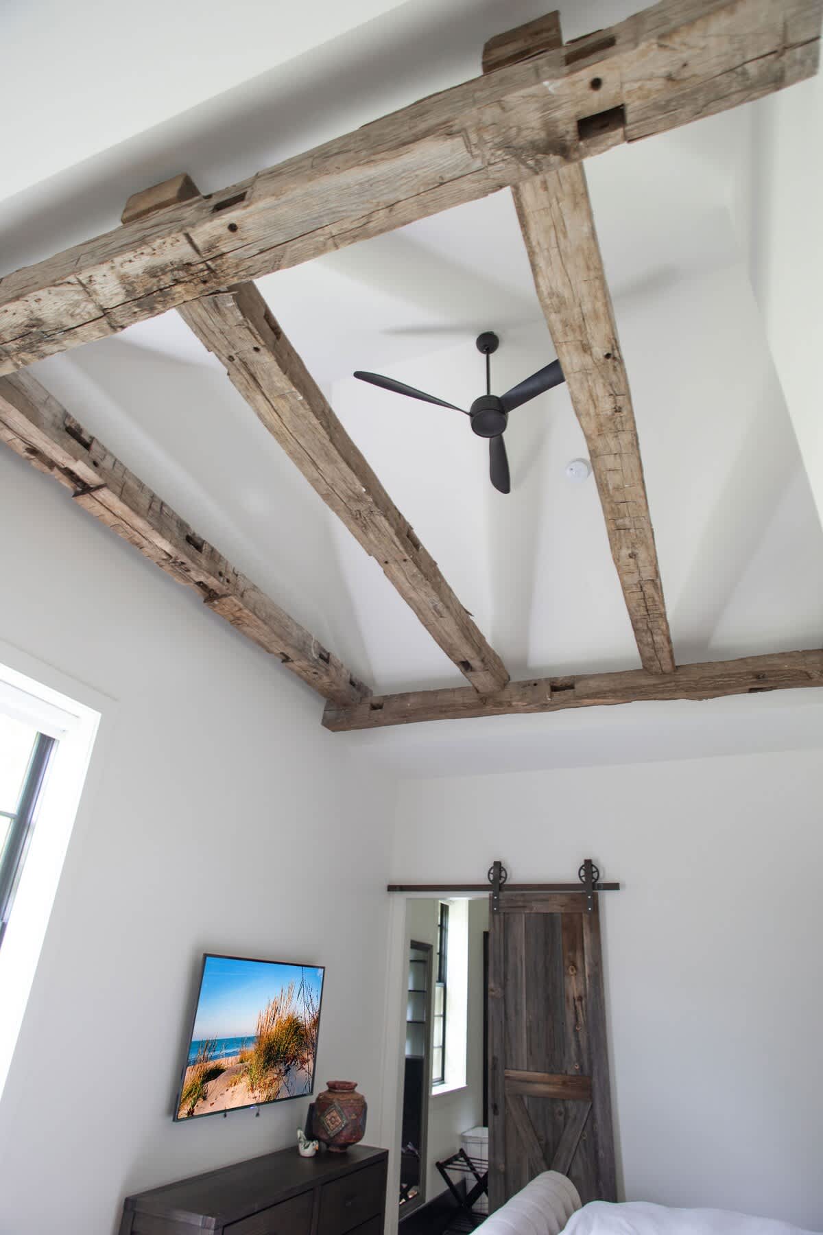 The bedroom showcases a striking white ceiling with prominent exposed wooden beams and a black ceiling fan. A wall-mounted television is positioned above a dark wooden dresser, with a rustic barn door visible in the corner.