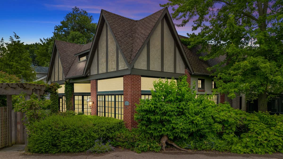The Tudor-style house exterior is illuminated at dusk, showcasing its steep gabled roof, brick and stucco facade, and large windows glowing with interior light, framed by dense green foliage.