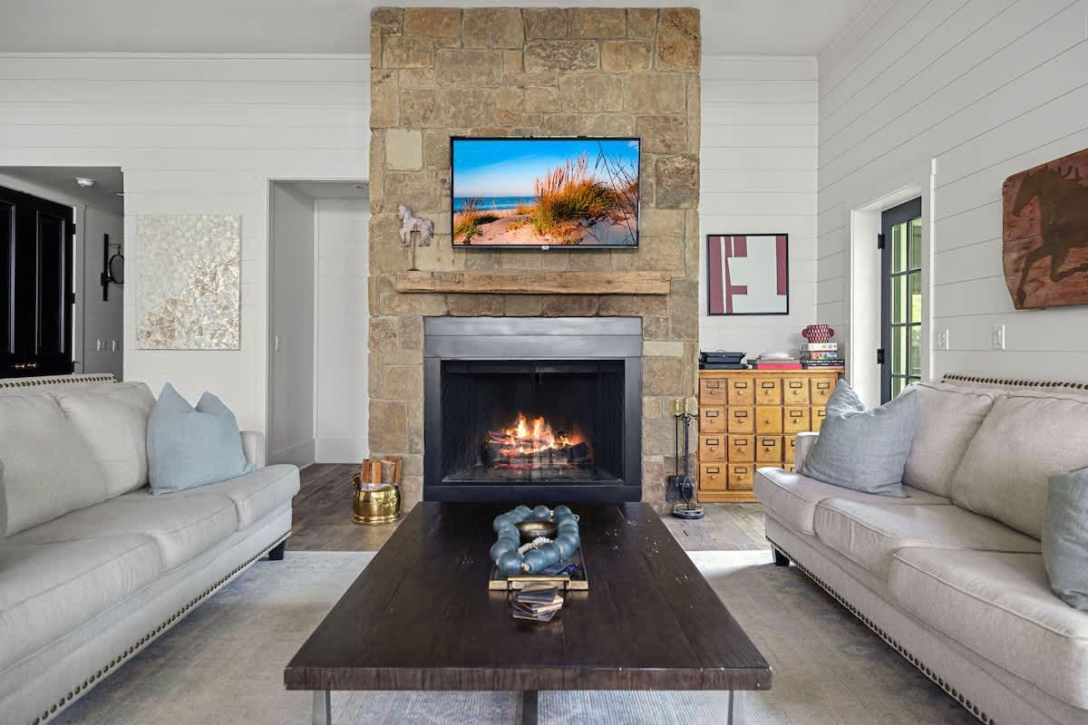 A cozy living room centered around a large stone fireplace with a wooden mantel, featuring a television mounted above. Two light-colored sofas with blue pillows face each other across a dark wood coffee table, and shiplap walls add texture.