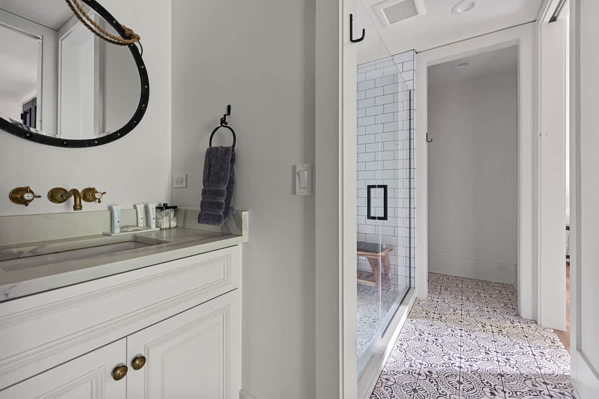 Modern bathroom featuring a white vanity with a marble countertop, a round black-framed mirror, and a walk-in shower with white subway tiles and a patterned floor. Gold fixtures add a touch of elegance.