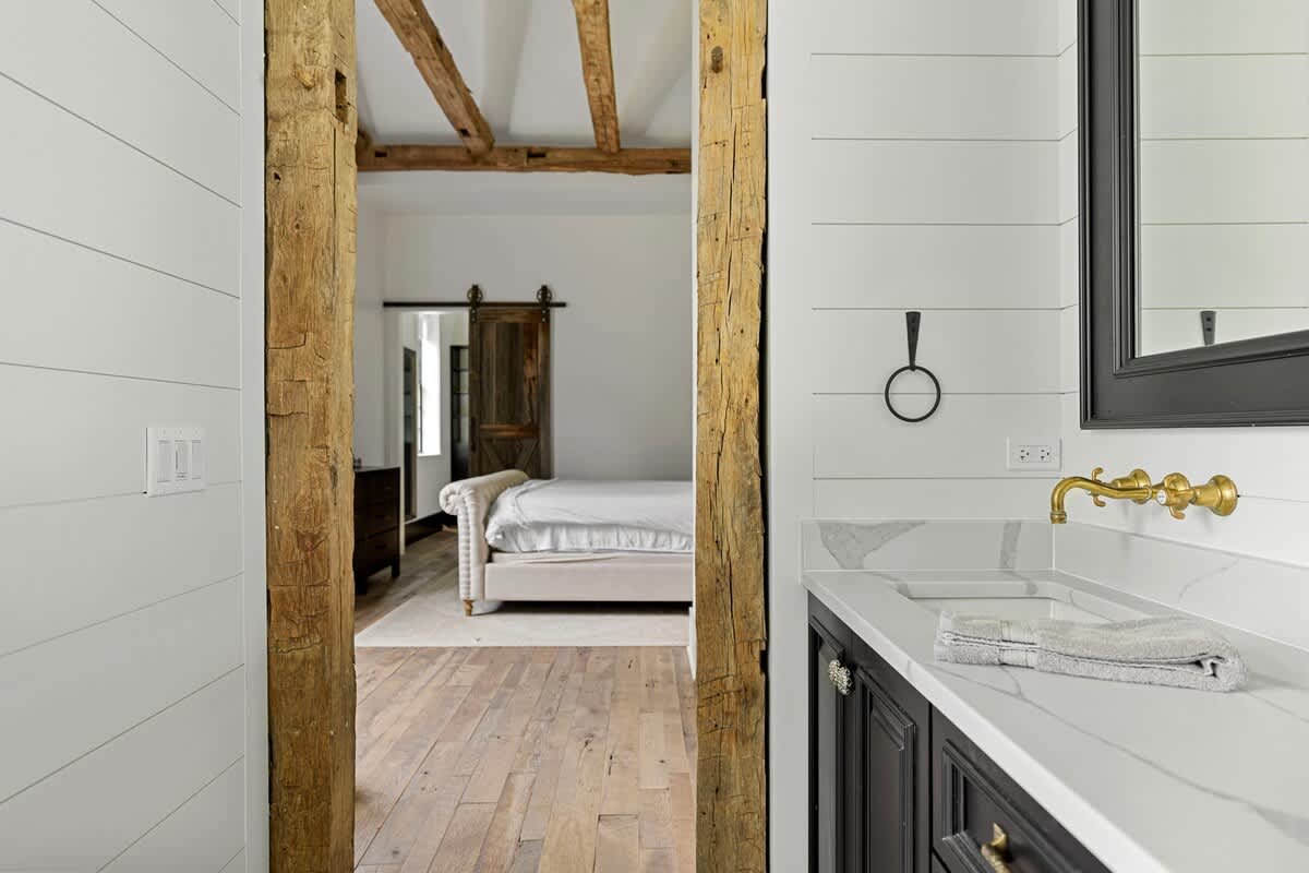 A modern bathroom vanity with a white countertop and gold faucet is seen from a doorway framed by rustic wooden beams, looking into a bedroom with an upholstered bed and a wooden barn door. White shiplap walls line the bathroom.