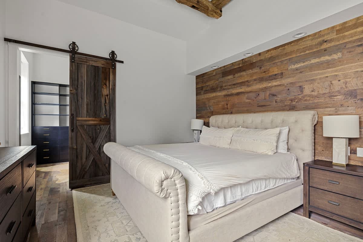 A cozy bedroom features a large, light-colored upholstered bed with a tufted headboard, set against a rustic wooden plank accent wall. A dark wooden barn door slides open to reveal a walk-in closet, with exposed wooden beams on the ceiling.