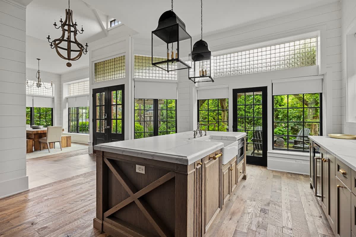 A spacious kitchen features a large wooden island with a white marble countertop and farmhouse sink, complemented by black pendant lights and white shiplap walls. Expansive black-framed windows offer views of the lush outdoors, with a dining area visible in the background.