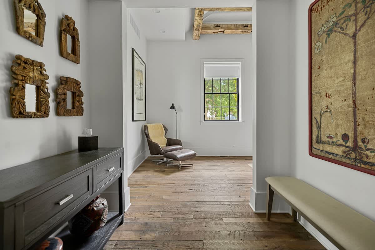 Inviting hallway with a dark console table adorned with decorative items and a wall featuring three ornate wooden mirrors. The hallway leads to a bright room with a modern lounge chair and large window.