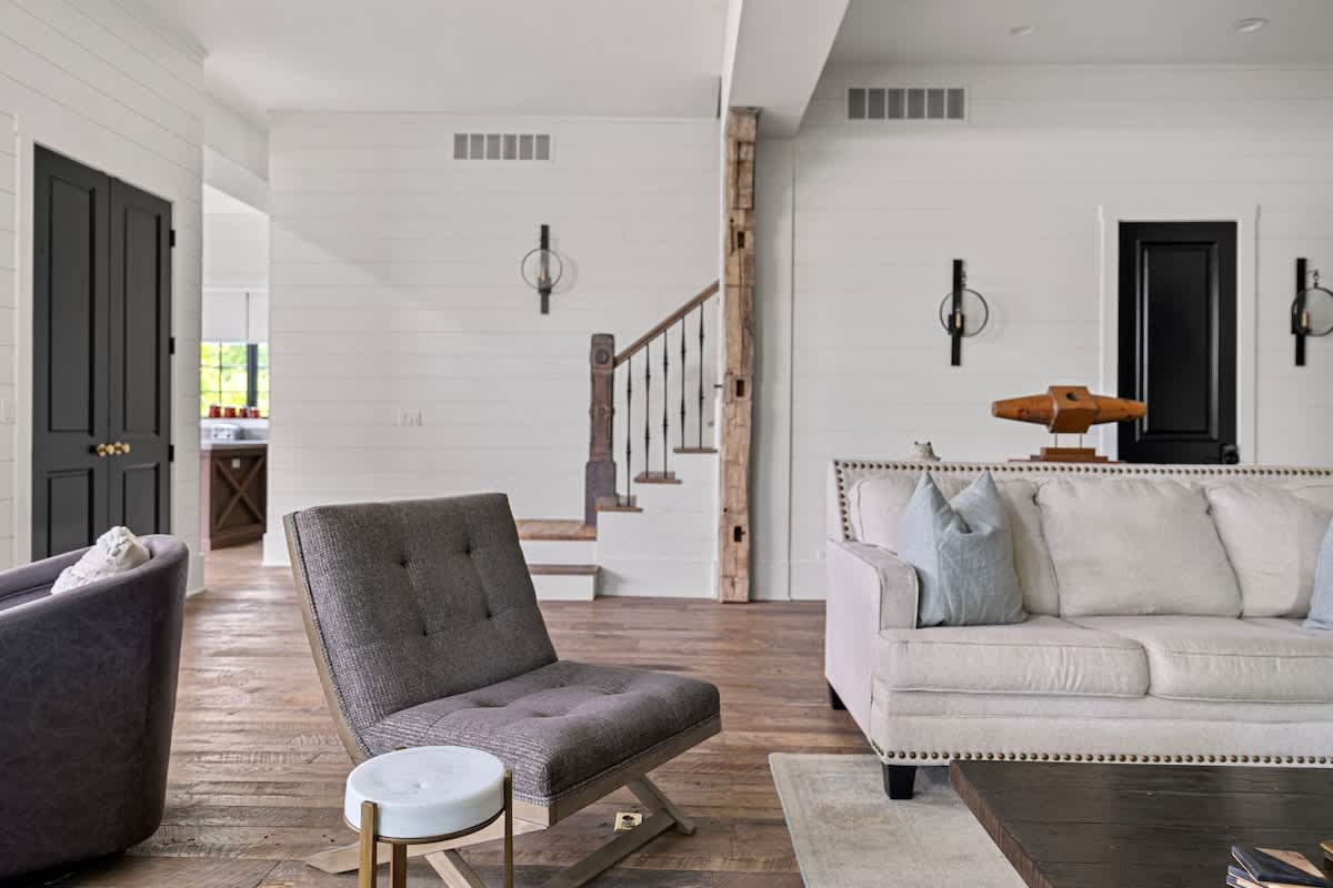 Stylish living room featuring a light-colored sofa, a gray upholstered chair, and dark paneled doors, with a wooden staircase visible in the background. Shiplap walls and rustic wall sconces add to the charm.