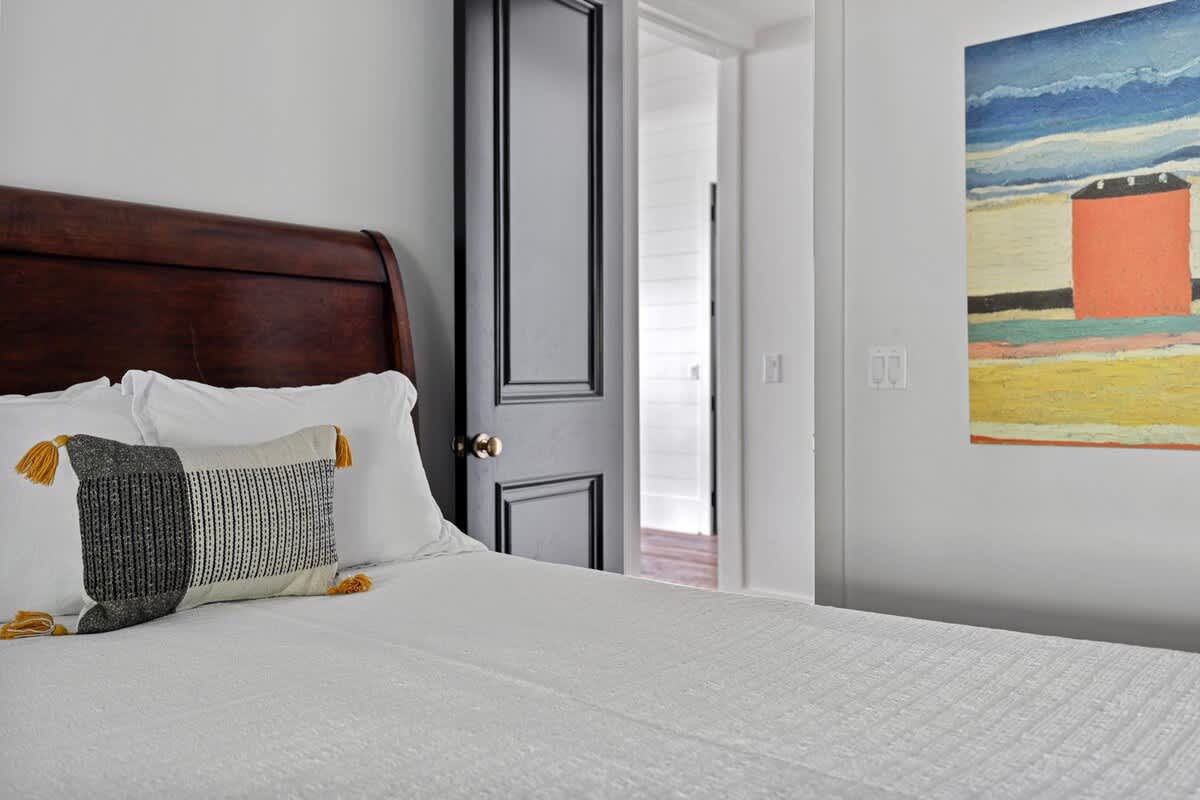 Cozy bedroom with a dark wooden headboard, white bedding, and a decorative pillow with black, white, and yellow accents. A black door is visible in the background, leading to another room.