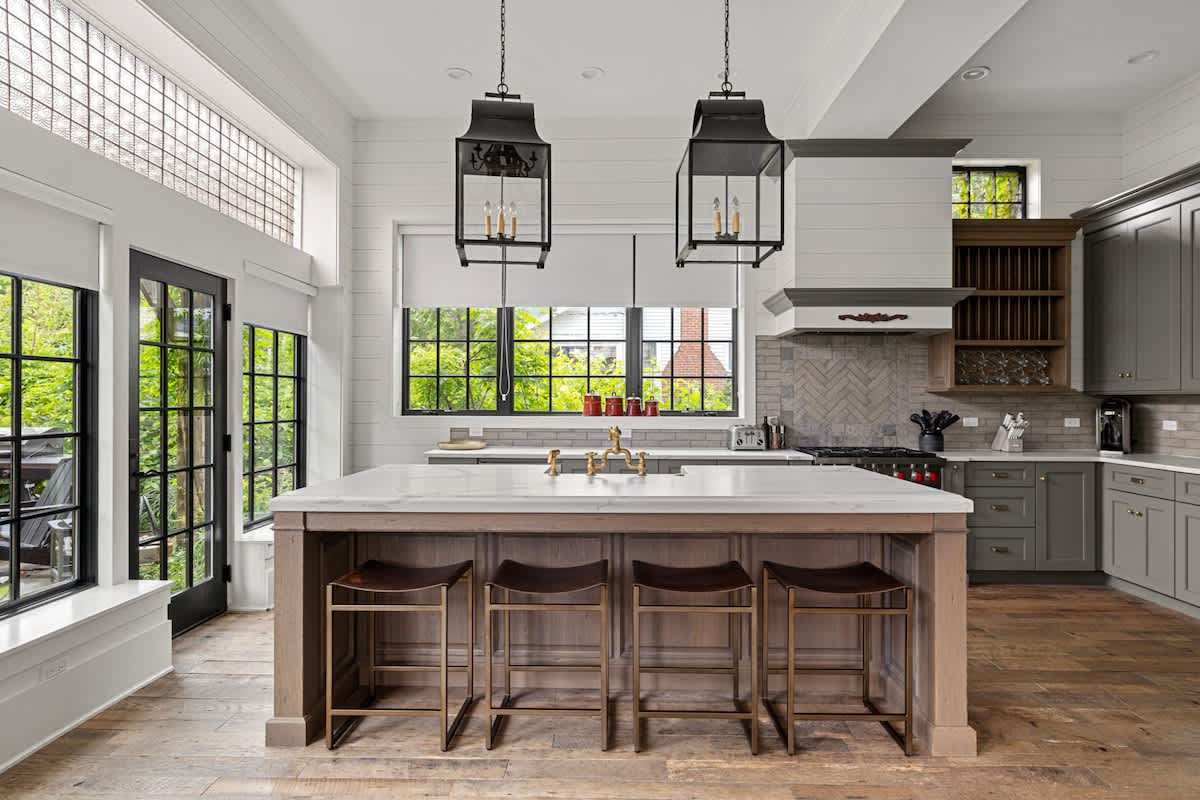 Spacious kitchen featuring a large white marble island with a dark wood base and four bar stools. Gray shaker cabinets line the walls, complemented by a light gray subway tile backsplash and black lantern pendant lights.