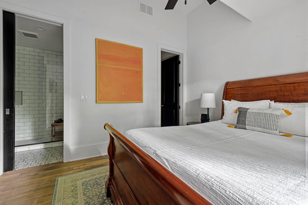 Bright bedroom featuring a large wooden sleigh bed with white bedding, an orange abstract painting on the wall, and an open doorway leading to a modern bathroom with white subway tiles.