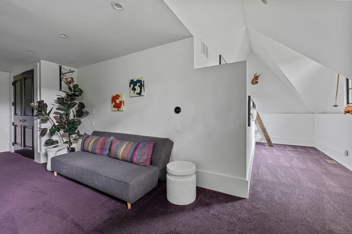 Lounge area with a gray sofa adorned with colorful striped pillows, abstract artwork on the wall, and purple carpet, leading into an attic-style play space.