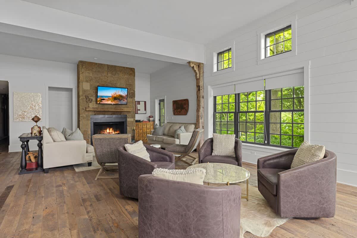 Expansive living area with a stone fireplace, multiple seating arrangements including sofas and swivel chairs, and large windows offering views of lush greenery. Rustic wooden beams add character to the bright space.
