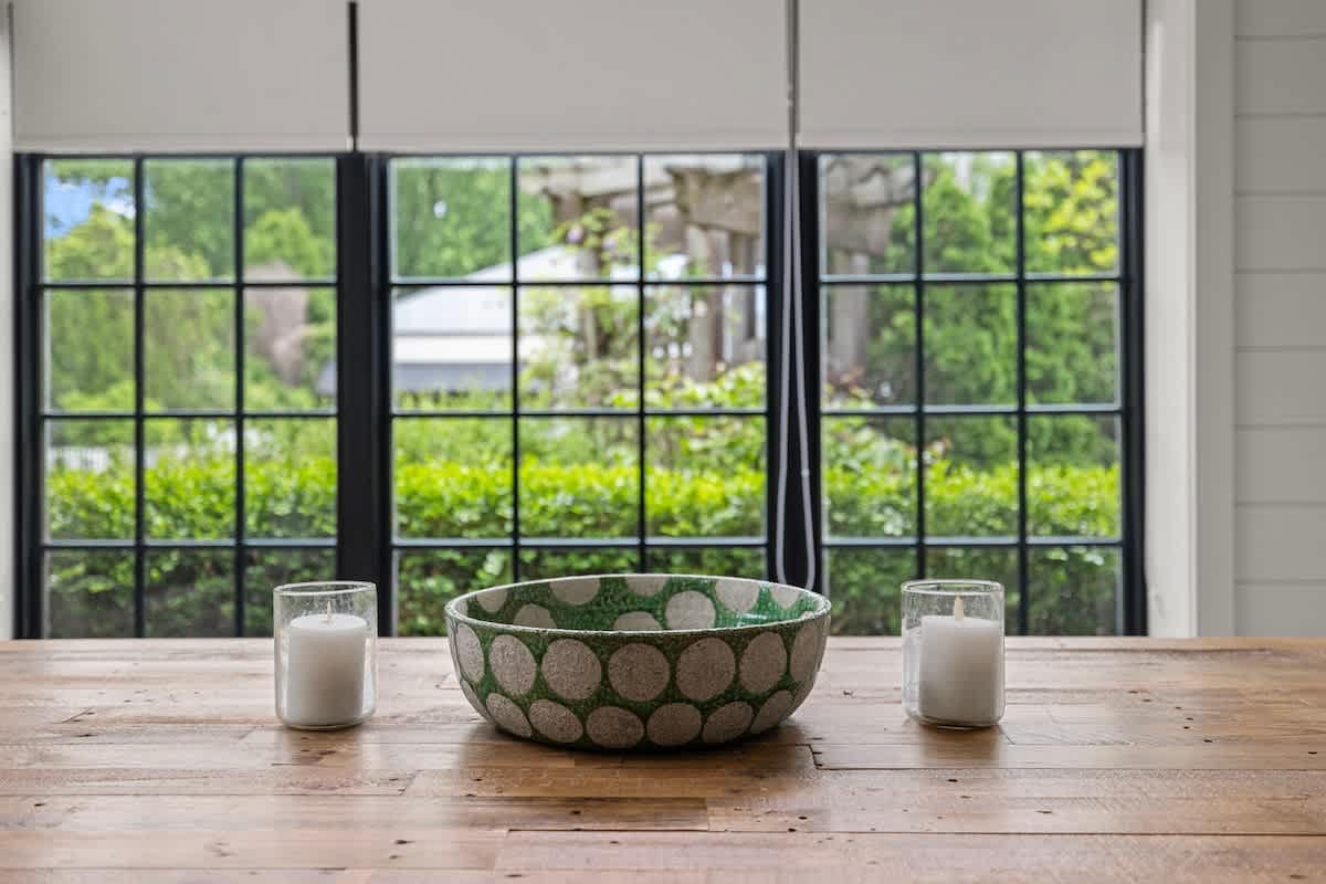 A rustic wooden dining table is adorned with a green and white patterned bowl and two white pillar candles, set against a backdrop of large black-framed windows overlooking lush green foliage.