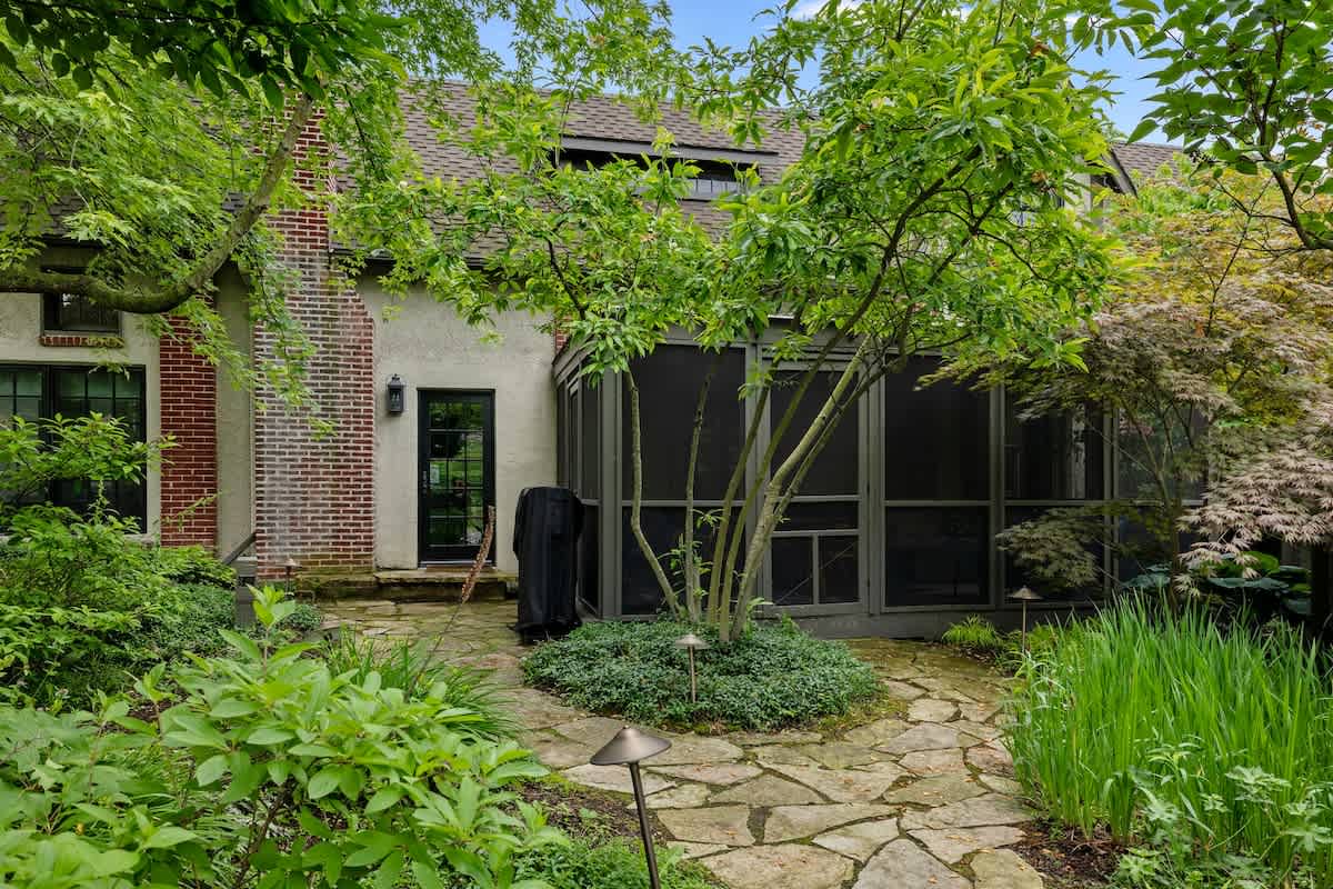 A flagstone path winds through a lush green garden, leading to a screened-in porch on the back of a brick and stucco house, surrounded by mature trees and shrubs.