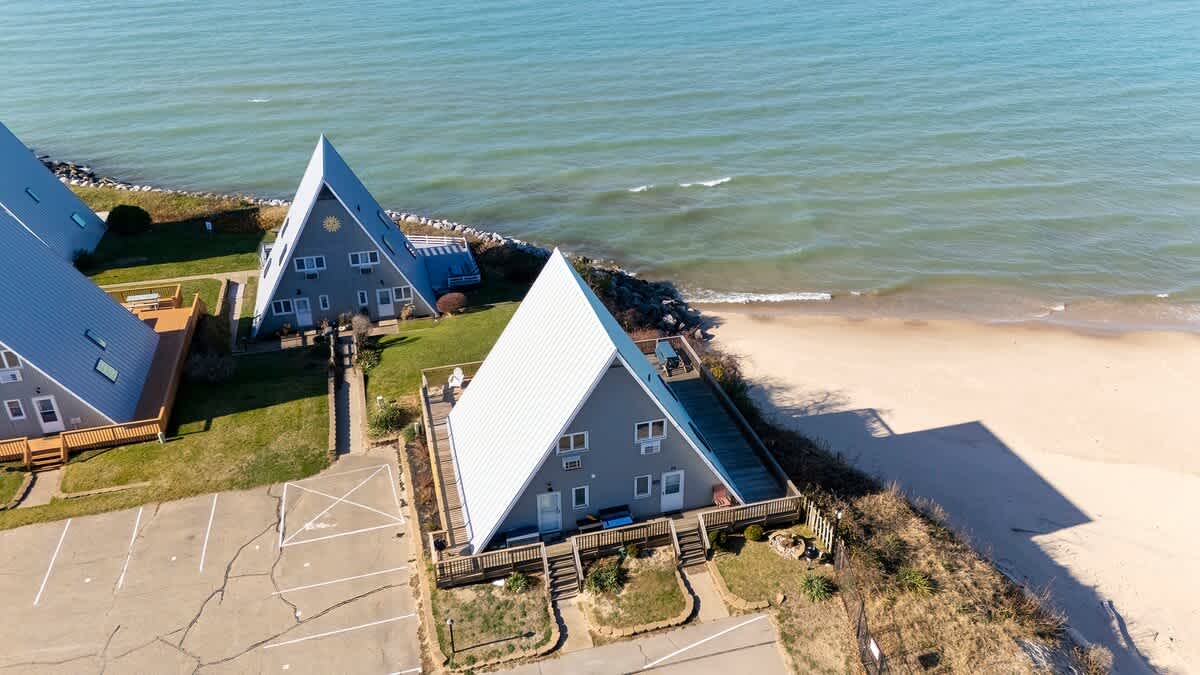 An aerial view showcases several A-frame chalets with gray siding and white roofs, situated along a sandy beach next to a calm blue lake. A parking area is visible in the foreground.