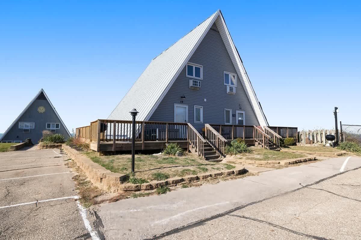 Front view of a gray A-frame chalet with a large wooden deck, stairs leading up to the entrance, and a paved parking area in the foreground. Another similar chalet is visible in the distance.