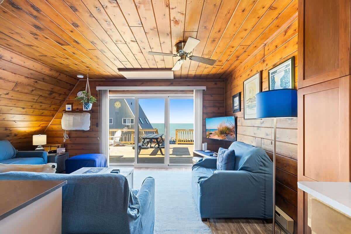 Bright living room with extensive wood paneling on walls and ceiling, furnished with blue seating, a ceiling fan, and a large sliding glass door providing a direct view of the outdoor deck and the water beyond. Artwork adorns the walls.