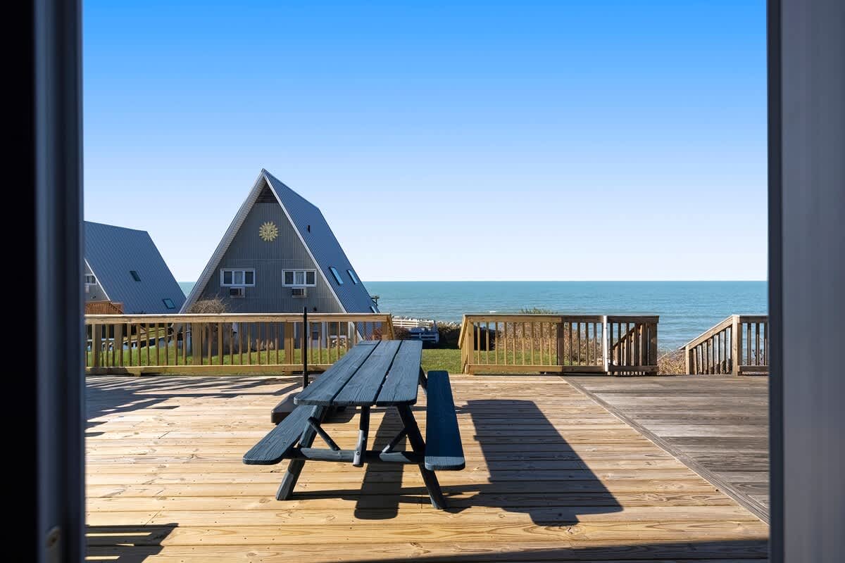 This view from inside a doorway looks out onto a large wooden deck with a blue picnic table, offering a panoramic view of the lake and a nearby A-frame chalet under a clear blue sky.
