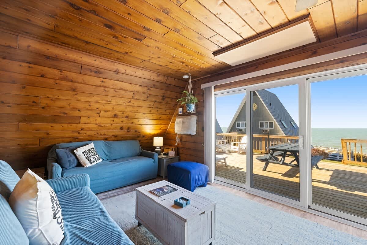 Living room with extensive wood paneling on walls and ceiling, featuring a blue sofa bed and another blue sofa, a light-colored coffee table, and a large sliding glass door opening to a deck with a view of another chalet and the water.