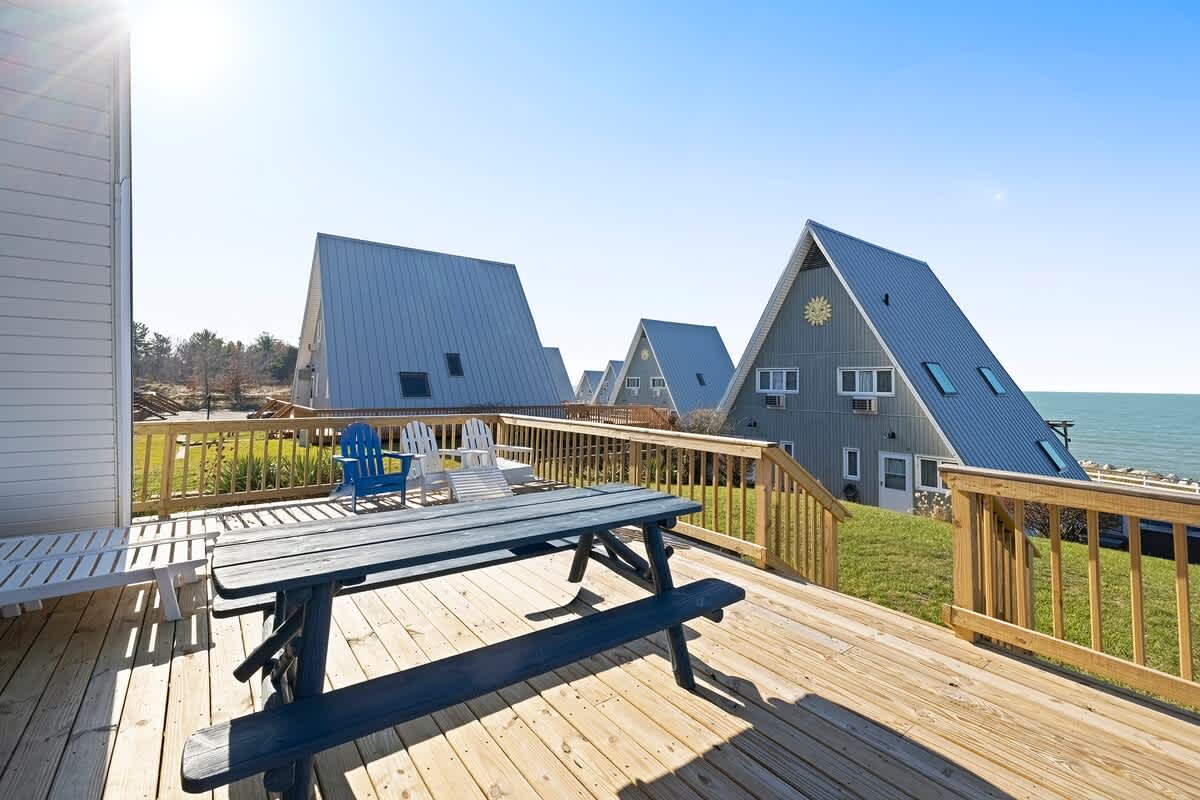 A sunny wooden deck features a blue picnic table and several Adirondack chairs, with multiple A-frame chalets visible in the background and the lake beyond. The deck is surrounded by wooden railings and green grass.