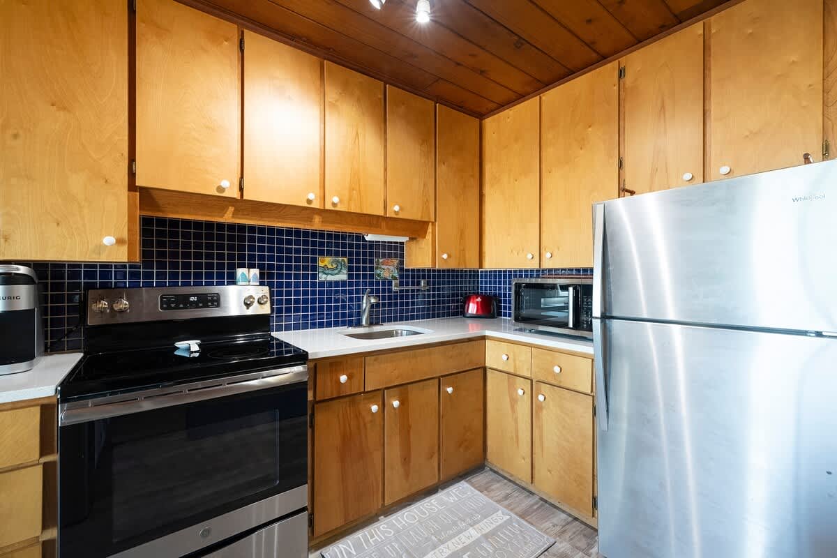 Well-equipped kitchen with light wood cabinets, a vibrant blue tiled backsplash, and stainless steel appliances, including a large refrigerator, electric range, and microwave, set against wood-paneled walls and ceiling.