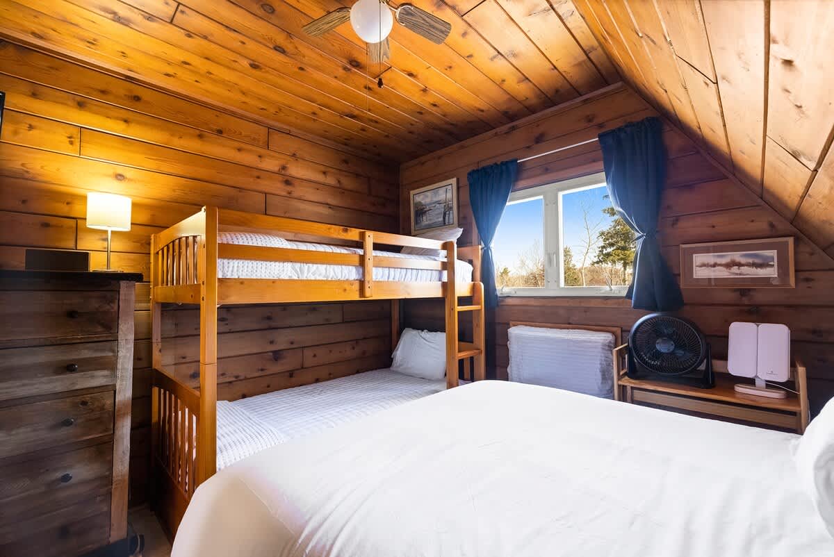 This bedroom, with its rustic wood-paneled walls and ceiling, includes a queen bed and a wooden bunk bed, with blue curtains framing a window that provides natural light. A ceiling fan and a small fan are also visible.